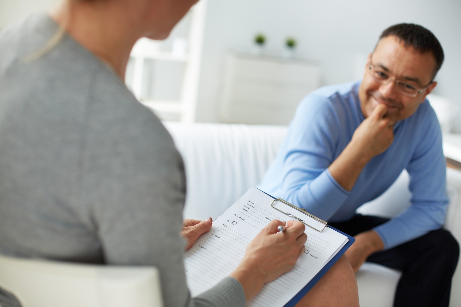 A therapist takes notes on a clipboard while engaging in a conversation with a thoughtful male client during a counseling session.