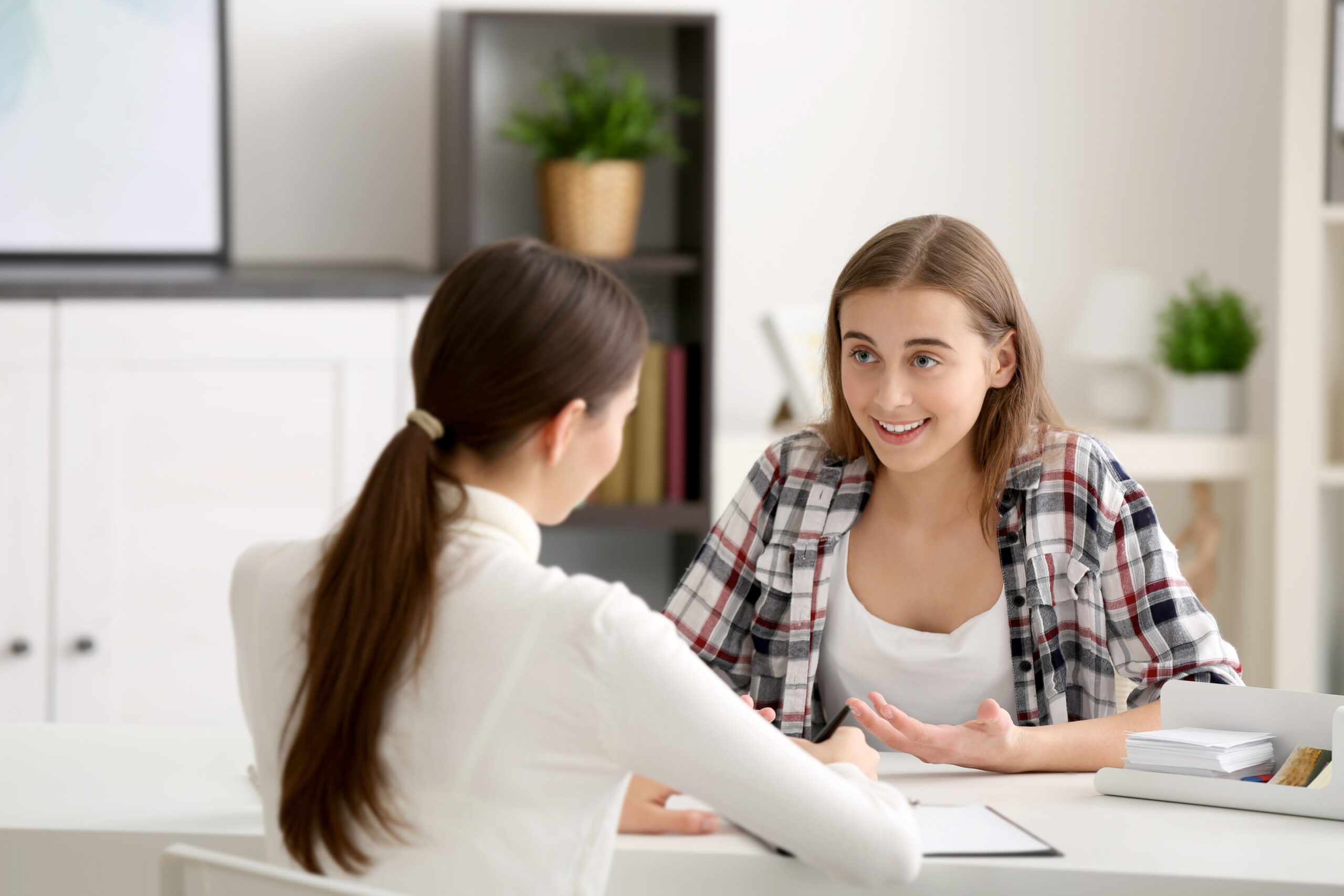 Two young women engage in a conversation at a desk, with one smiling and gesturing animatedly while the other listens attentively. The setting is bright and modern.